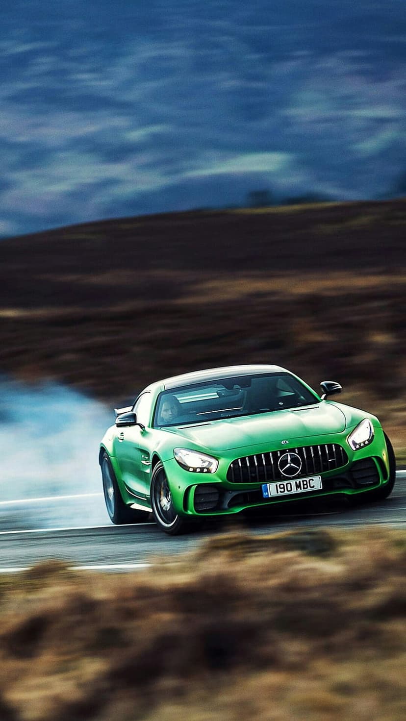 Green Mercedes-AMG GT car drifts sideways on a mountain road with white tire smoke against rolling hills.