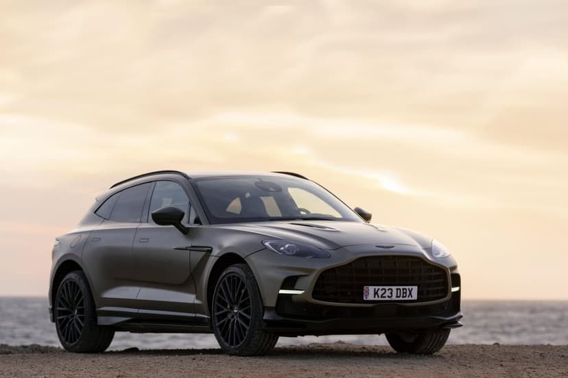 Bronze Aston Martin DBX707 luxury SUV parked on a rugged sandy beach under a hazy golden sunset and ocean horizon.