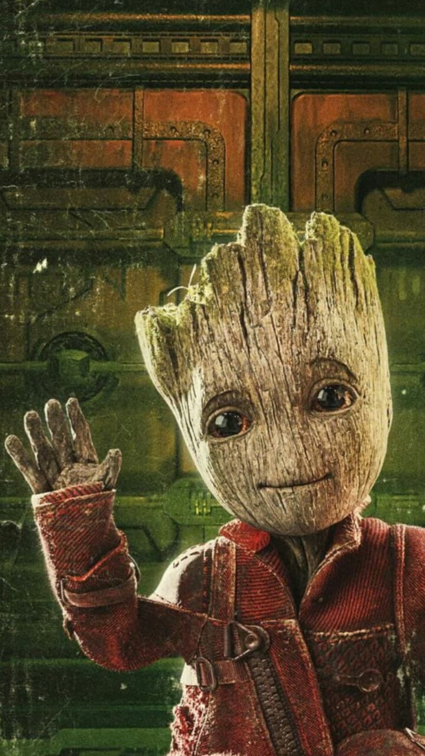 Baby Groot in a red zippered jacket waves his hand while standing against a rusted, metallic industrial wall.