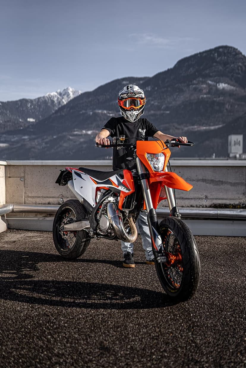 Motorcycle rider in orange gear poses with a supermoto bike before snow-covered mountains and a clear blue sky.