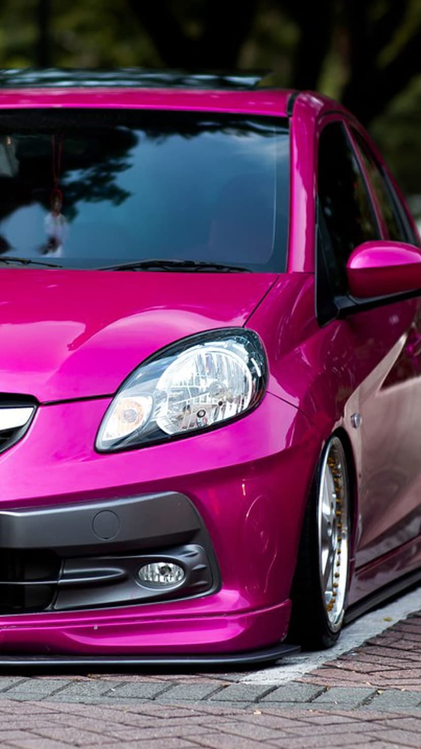 Metallic pink car with gold rims and tinted windows parked on a brick road under soft daylight reflections.