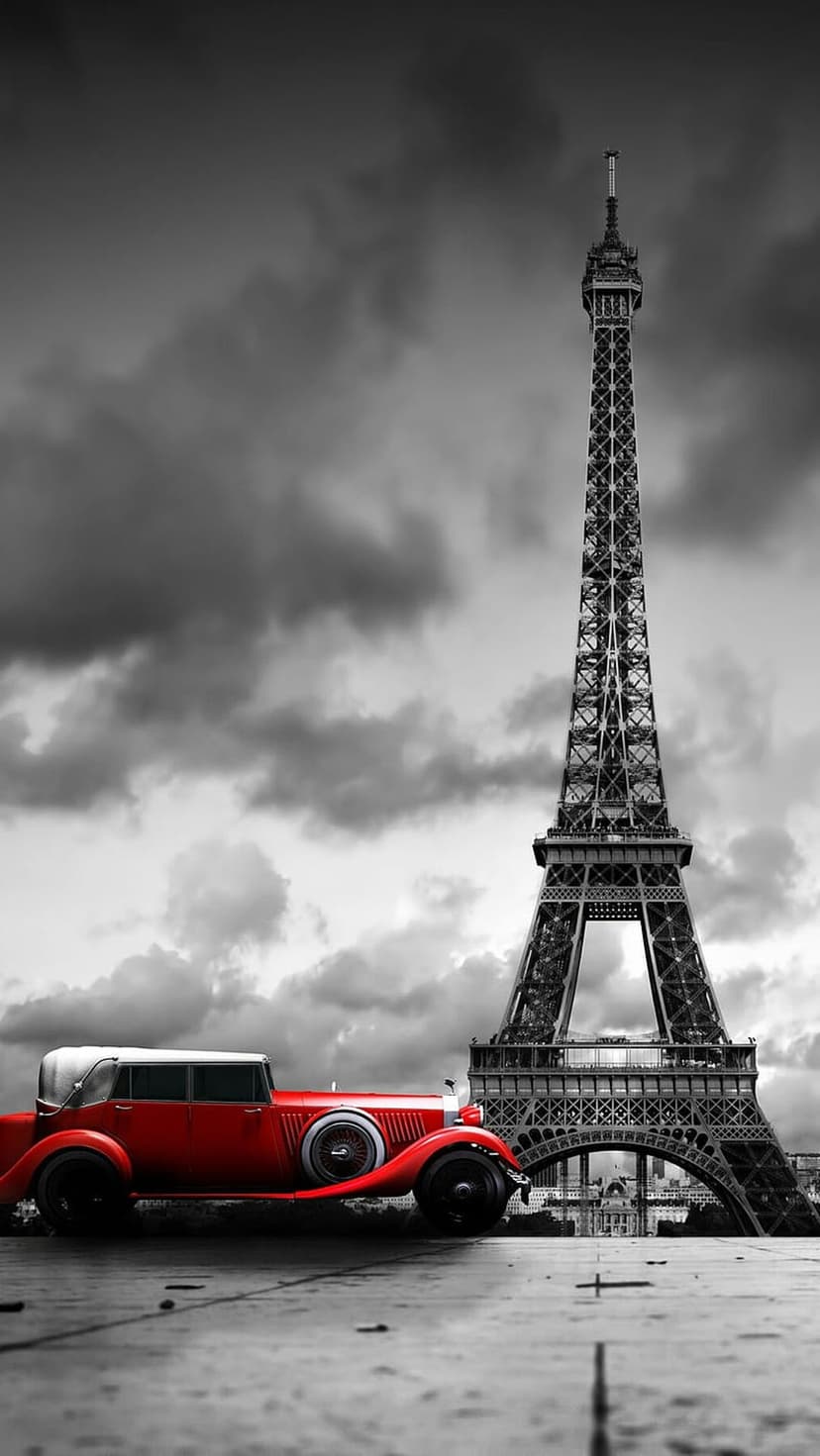 Red vintage car parked on a Parisian street with the Eiffel Tower and a moody monochrome sky in the background.