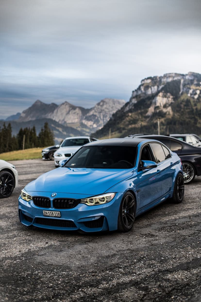 Blue BMW M4 parked on a gravel trail with a massive mountain range under a cloudy sky during an outdoor car meet.