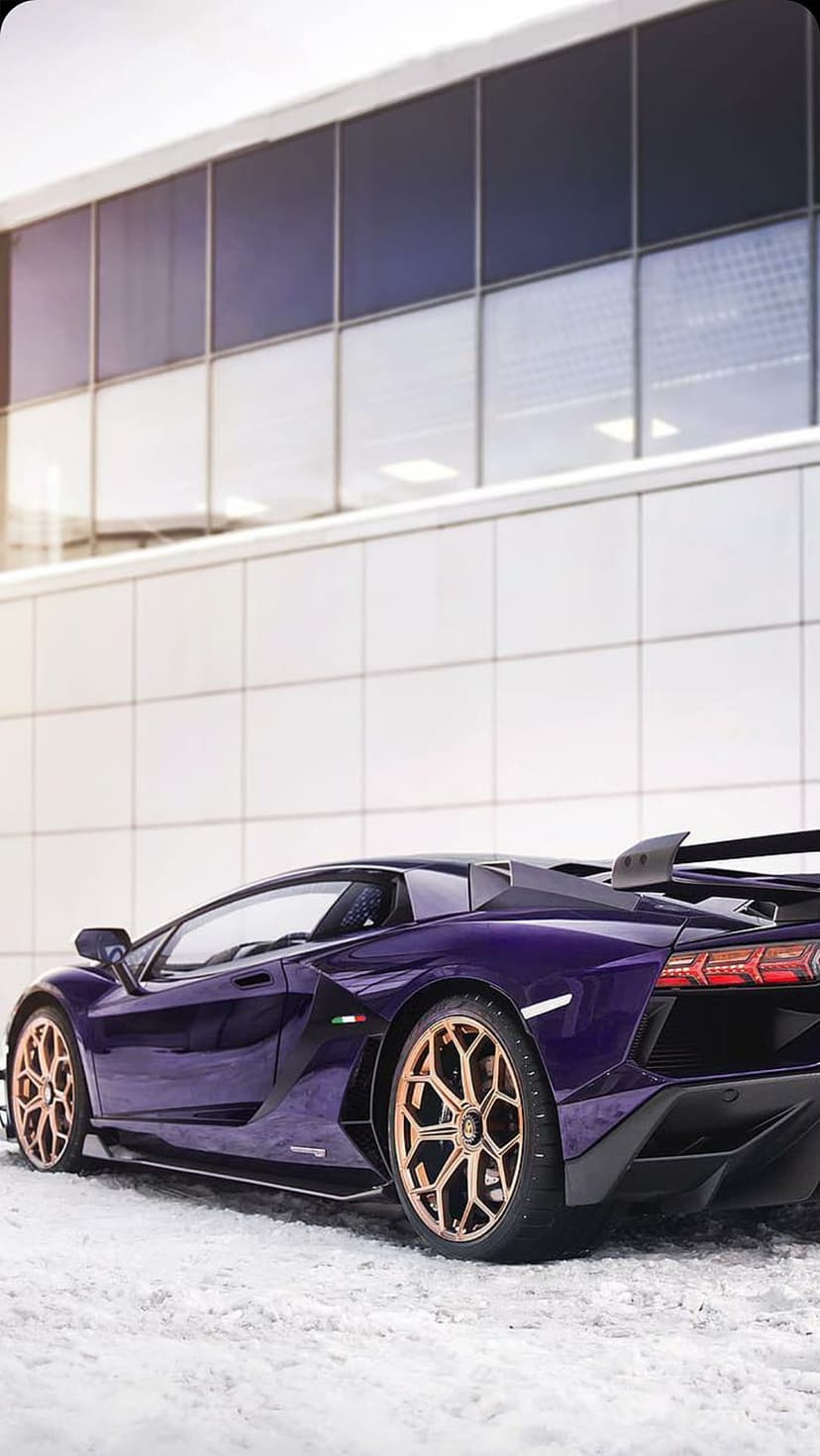 Purple Lamborghini Aventador with gold wheels parked on snow by a white tiled building in a portrait car wallpaper.