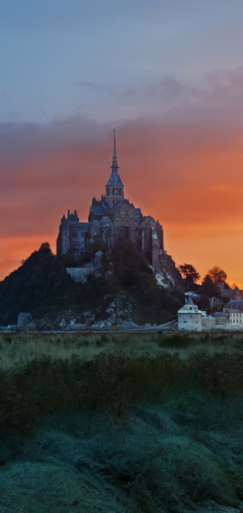 Mont Saint-Michel abbey silhouetted against a vibrant orange and red sunset sky with green tidal marshes below.