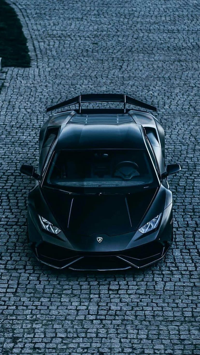 Black Lamborghini Huracan with a rear wing parked on a cobblestone street seen from a top-down aerial perspective.