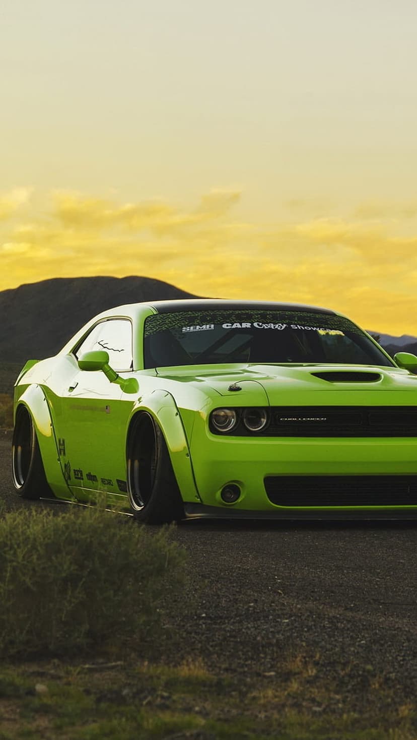 Lime green Dodge Challenger widebody on a gravel road under a yellow sunset sky with distant rolling hills.