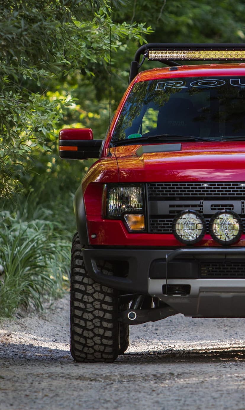 Red Ford Raptor truck with roof rack and LED lights parked on a dirt road surrounded by green forest plants.