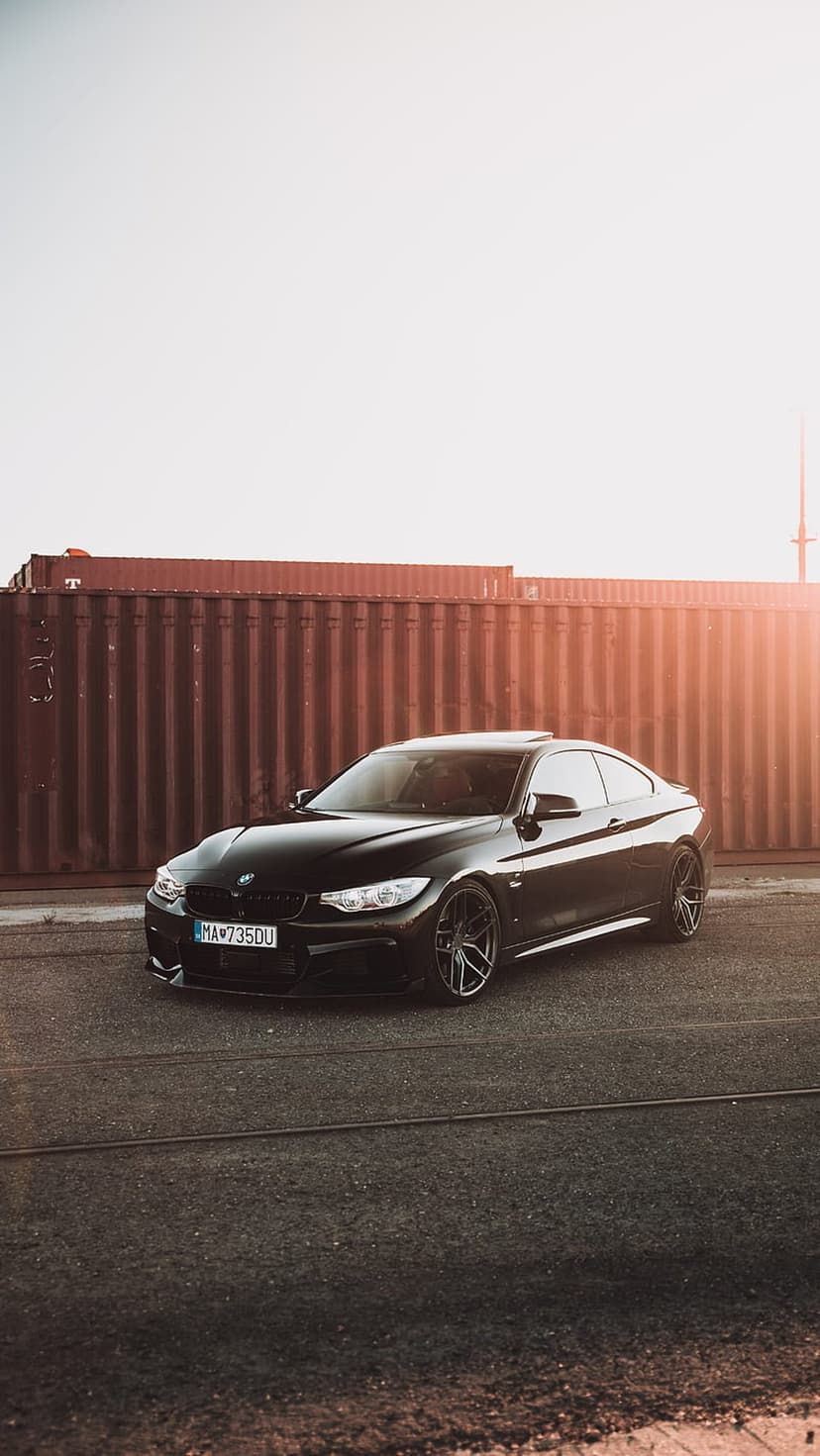 Black BMW 4 Series coupe parked by red shipping containers under golden sunlight on an asphalt lot.