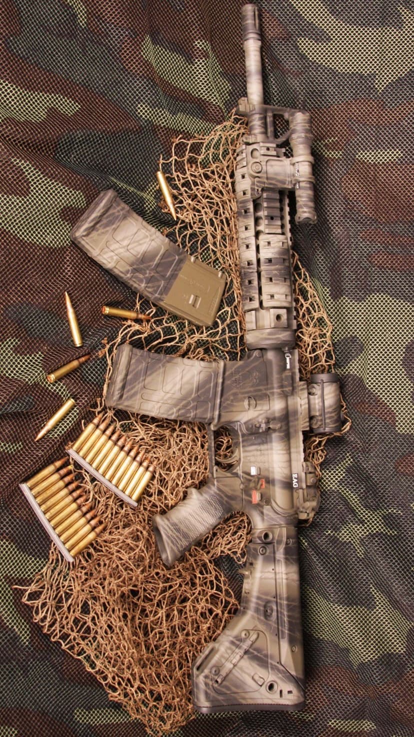 Tactical rifle with camouflage paint rests on mesh netting beside brass ammunition and several magazines.