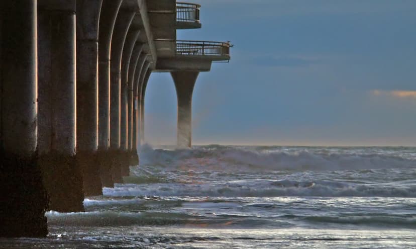 Massive concrete pier pylons endure crashing white ocean waves during a vibrant golden hour sunrise at sea.
