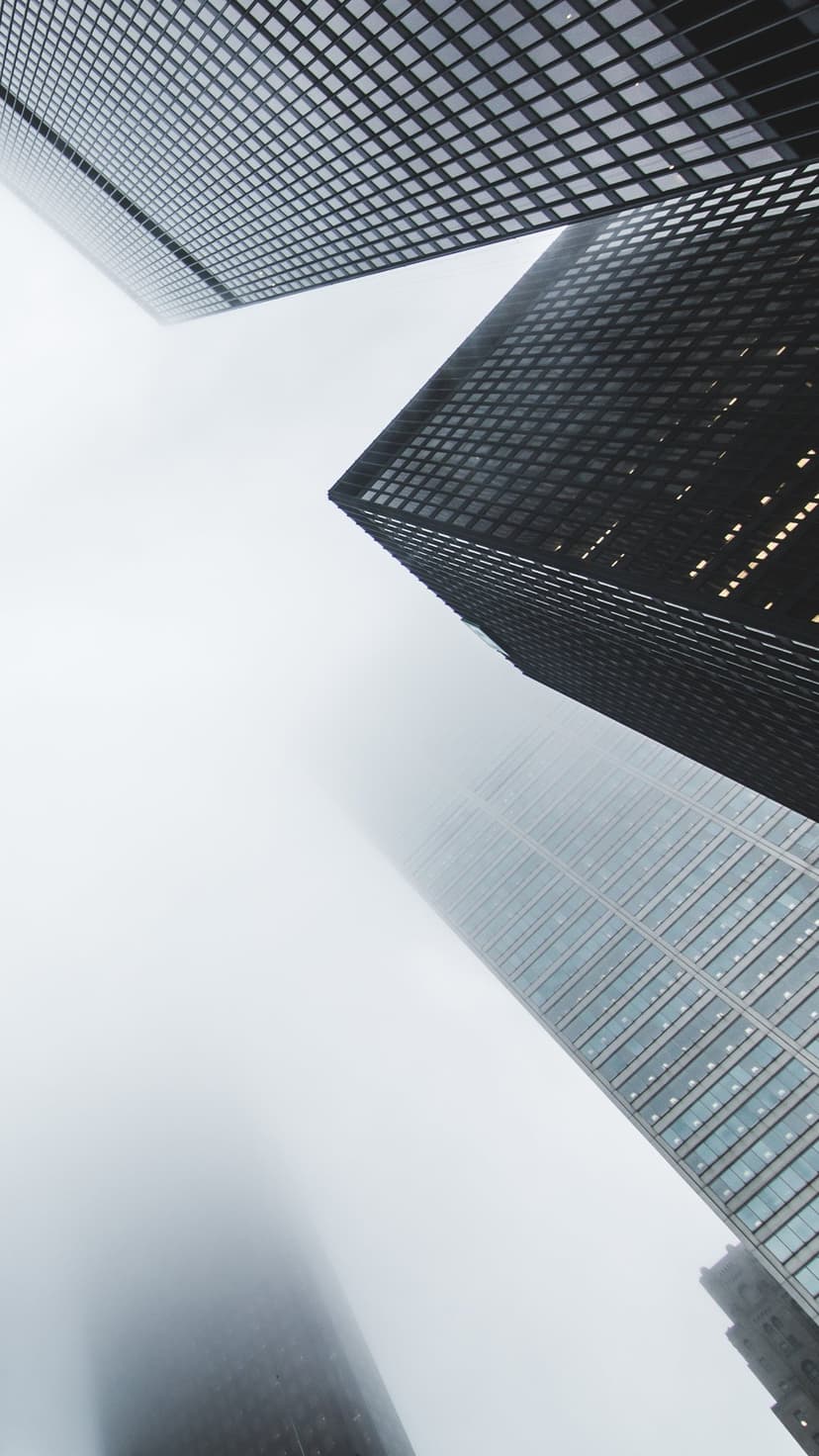 Skyscrapers vanishing into dense fog from an upward perspective with glass facades and glowing office lights.