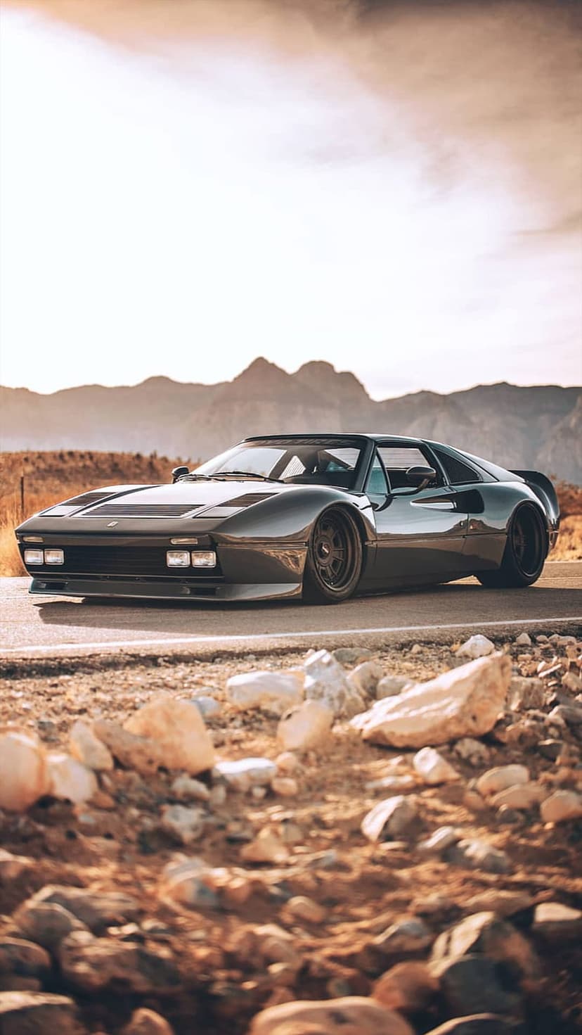 Ferrari 308 GTB with custom wheels parked on a rocky desert road during golden hour against mountain silhouettes.