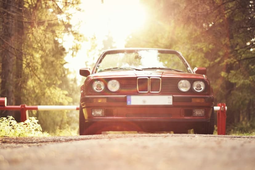 Red vintage BMW convertible parked on a gravel path during sunset with light rays filtering through green trees.