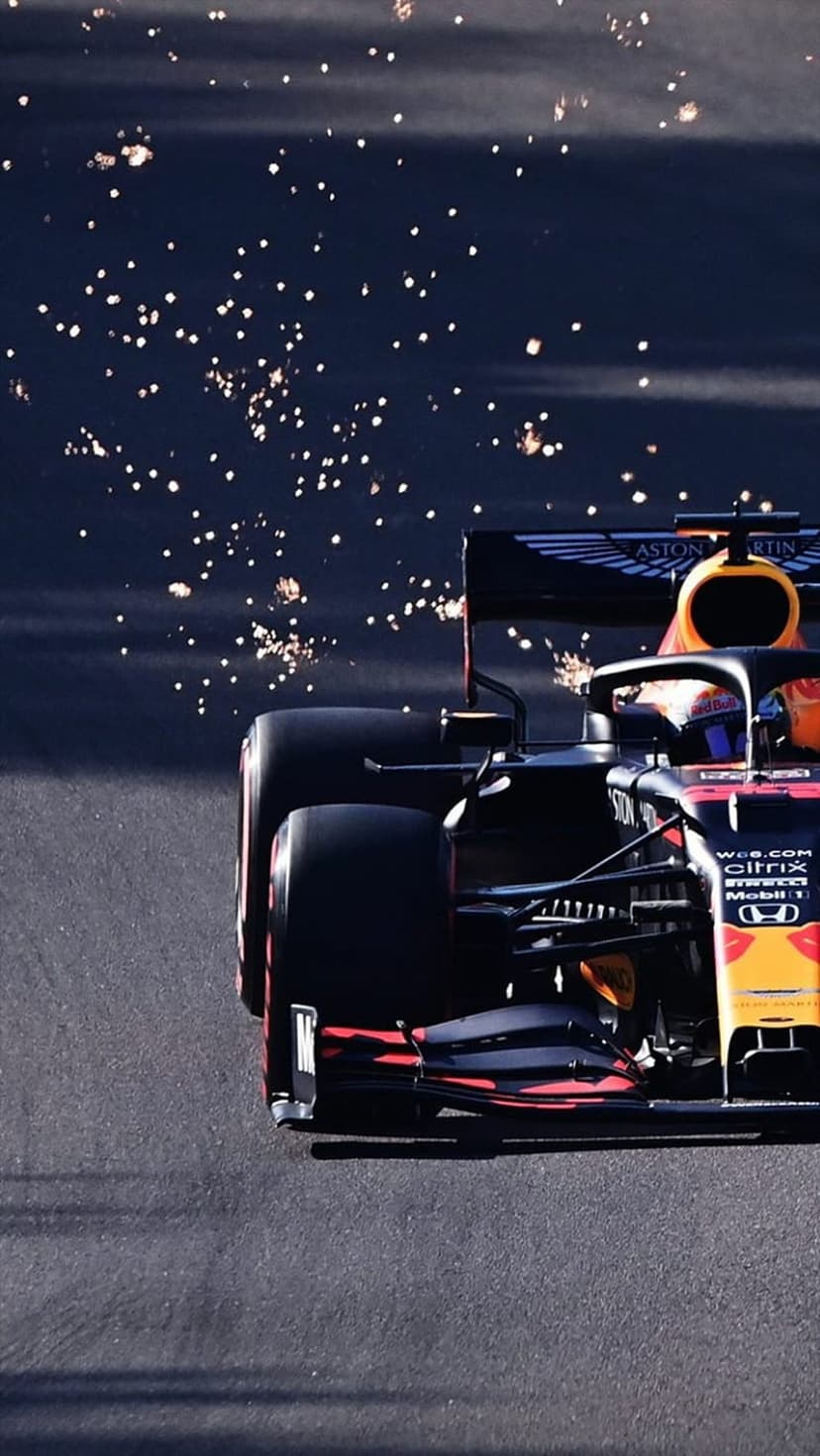 Red and black F1 car racing on a dark asphalt track with bright pyrotechnic sparks glowing in the background.