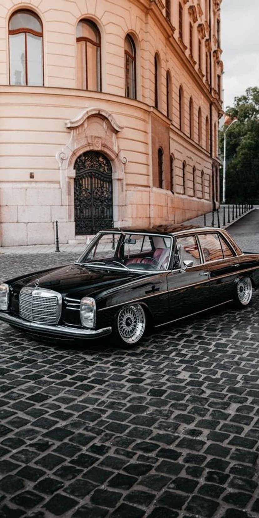 Black vintage Mercedes-Benz parked on a cobblestone street by a stone building with arched windows and iron gates.
