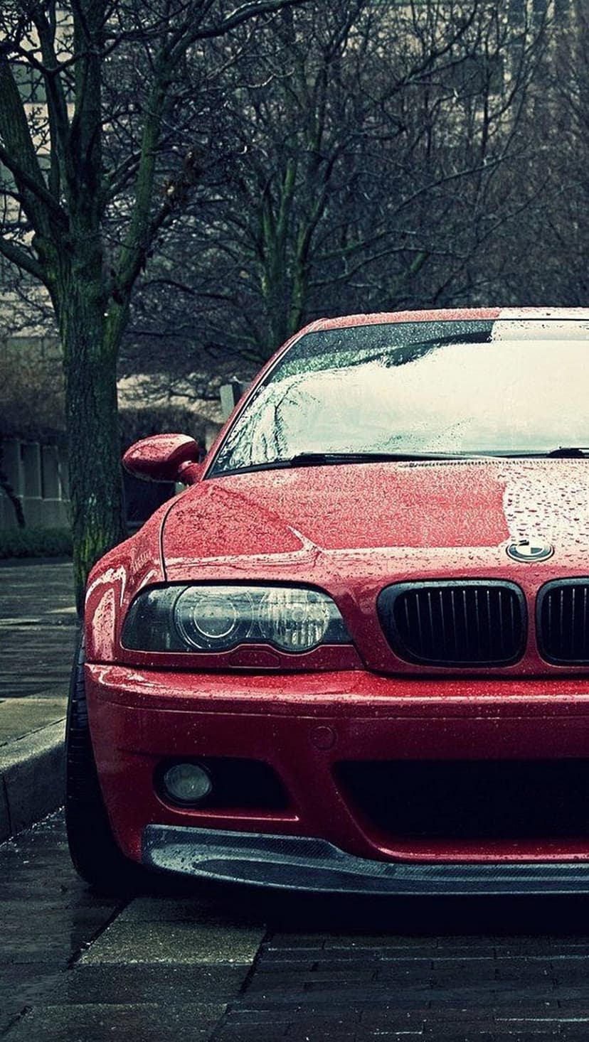 Red BMW M3 with glowing headlights parked on a wet street with water droplets on the hood during rainfall.