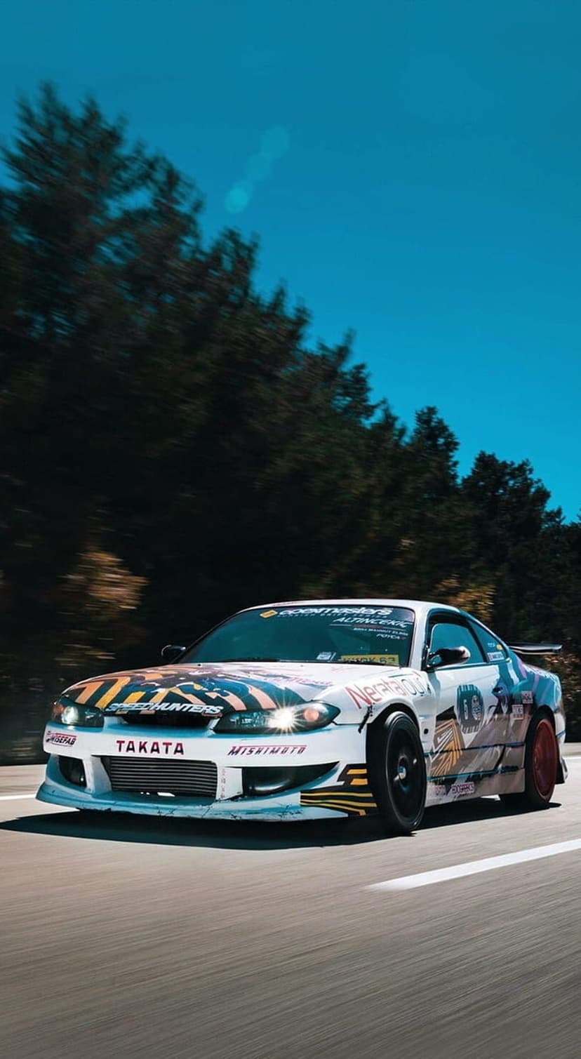 White sports car with Takata decals drifting through a corner against a blurred background of green trees.