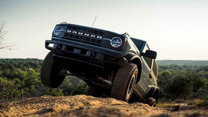 Ford Bronco off-roading on a sandy hill with one wheel lifted against a green valley and bright sky.