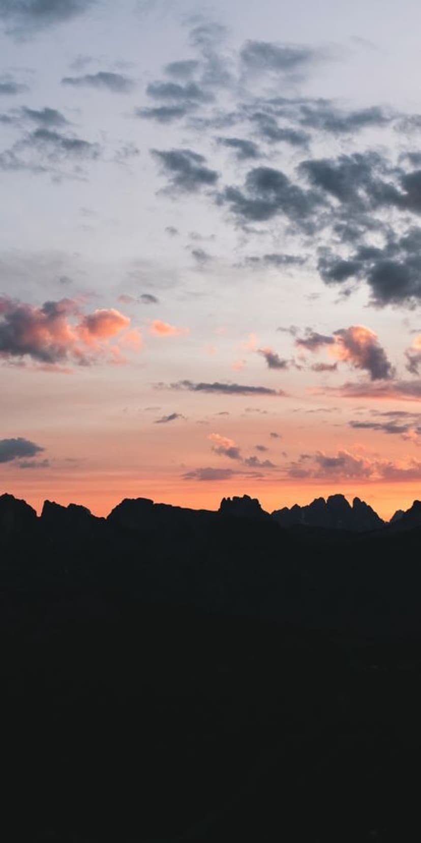 Jagged mountain range silhouettes under a vibrant orange and pink sunset sky with scattered grey clouds.