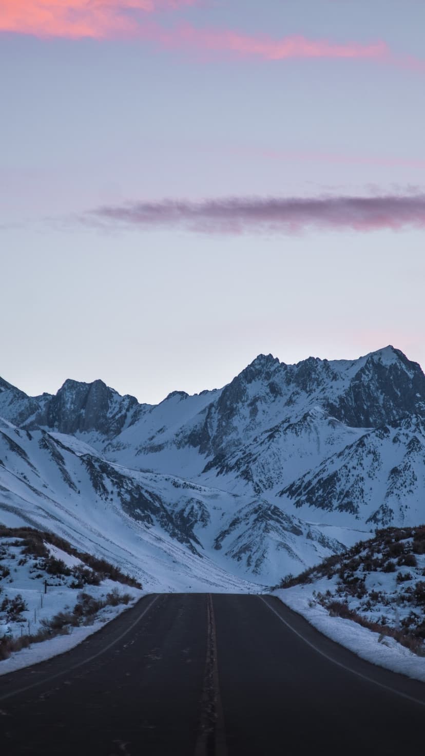 Snow-capped mountains loom over a long road under pink and purple twilight clouds in a vertical winter landscape.
