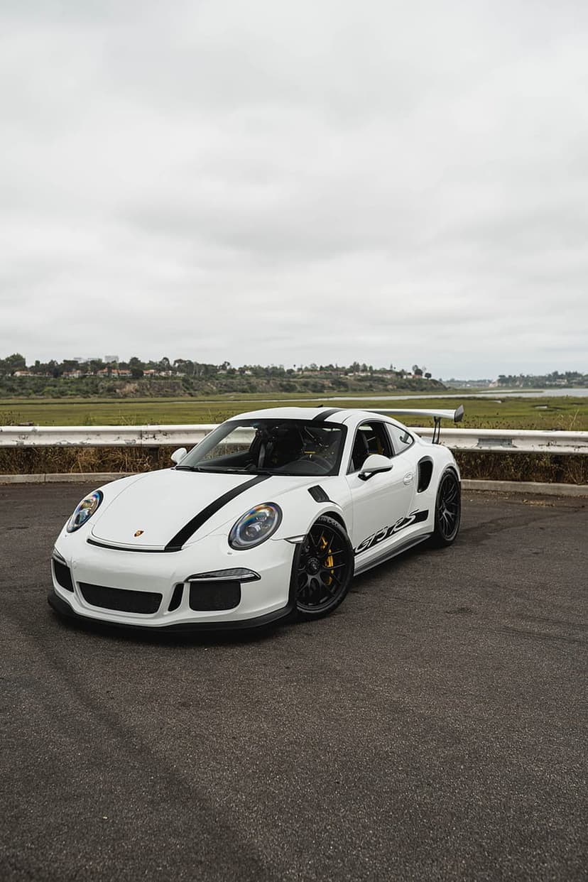 White Porsche 911 GT3 RS with black racing stripes parked on asphalt near a cloudy coastline with a large rear wing.