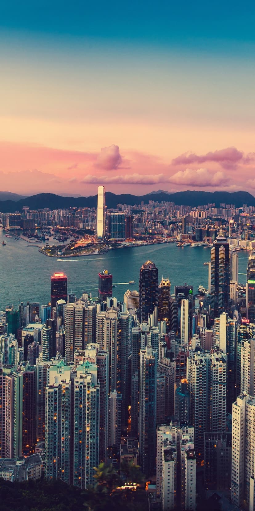 Glowing Hong Kong skyscrapers border a calm bay beneath a soft pink and blue twilight sky with mountain peaks.