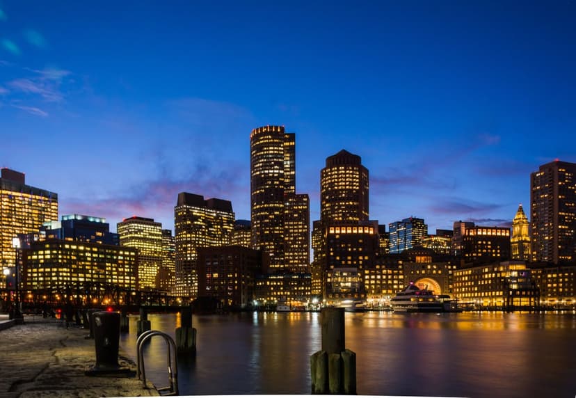 Illuminated Boston skyscrapers reflect in dark harbor water beneath a deep blue twilight sky during dusk.