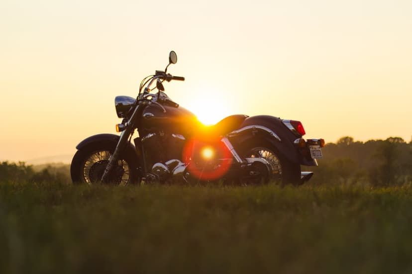 Motorcycle silhouette stands in a tall grass field at sunset with a warm golden lens flare and amber sky.