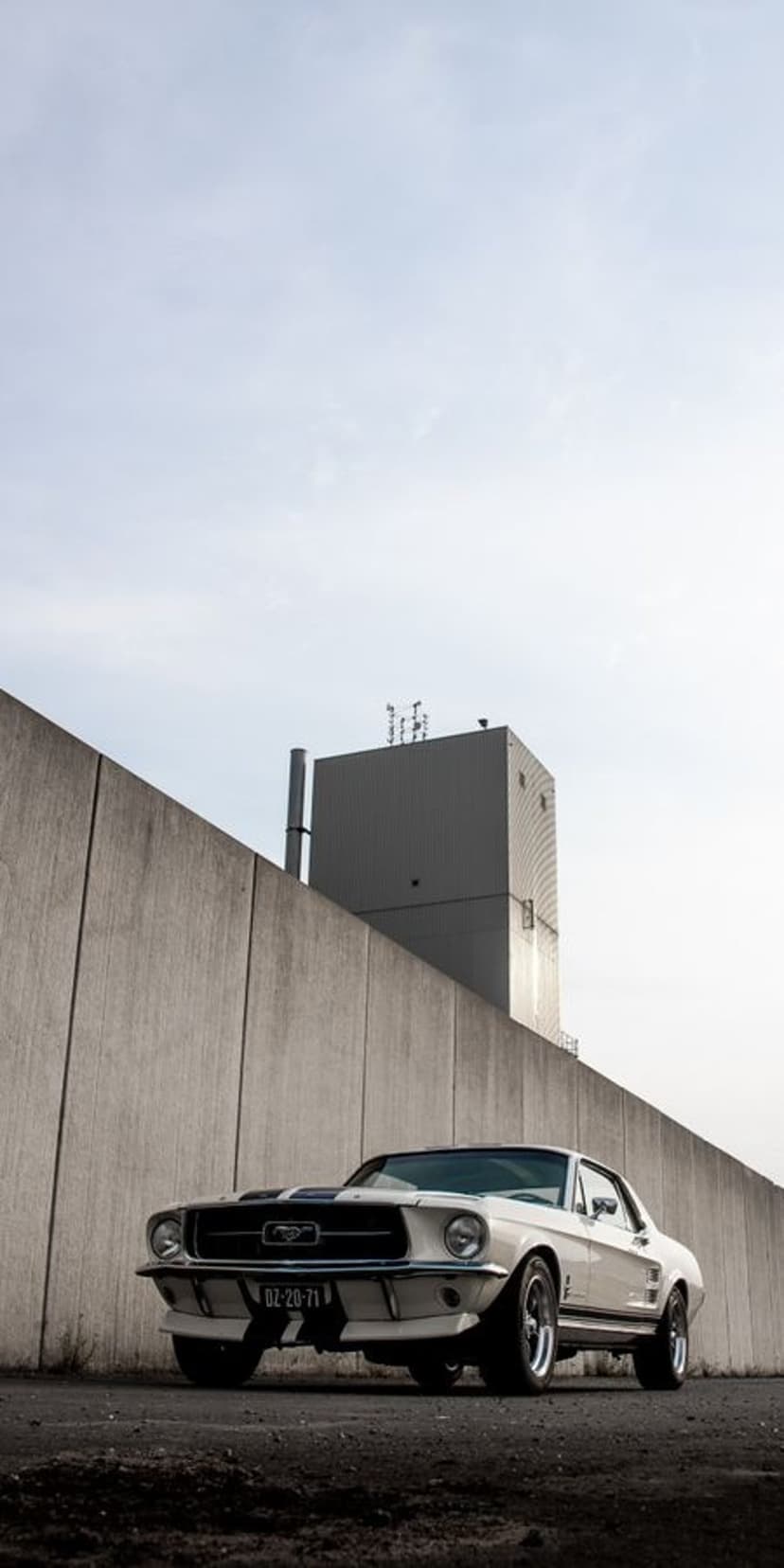 White Ford Mustang with black racing stripes parked by a concrete wall with a factory chimney in the distance.