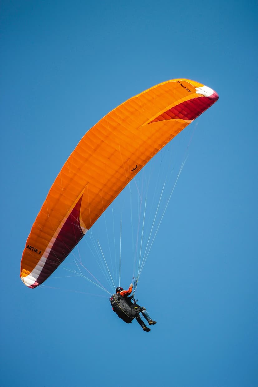 Paraglider in an orange jacket suspended under an orange and white wing against a clear, deep blue sky.