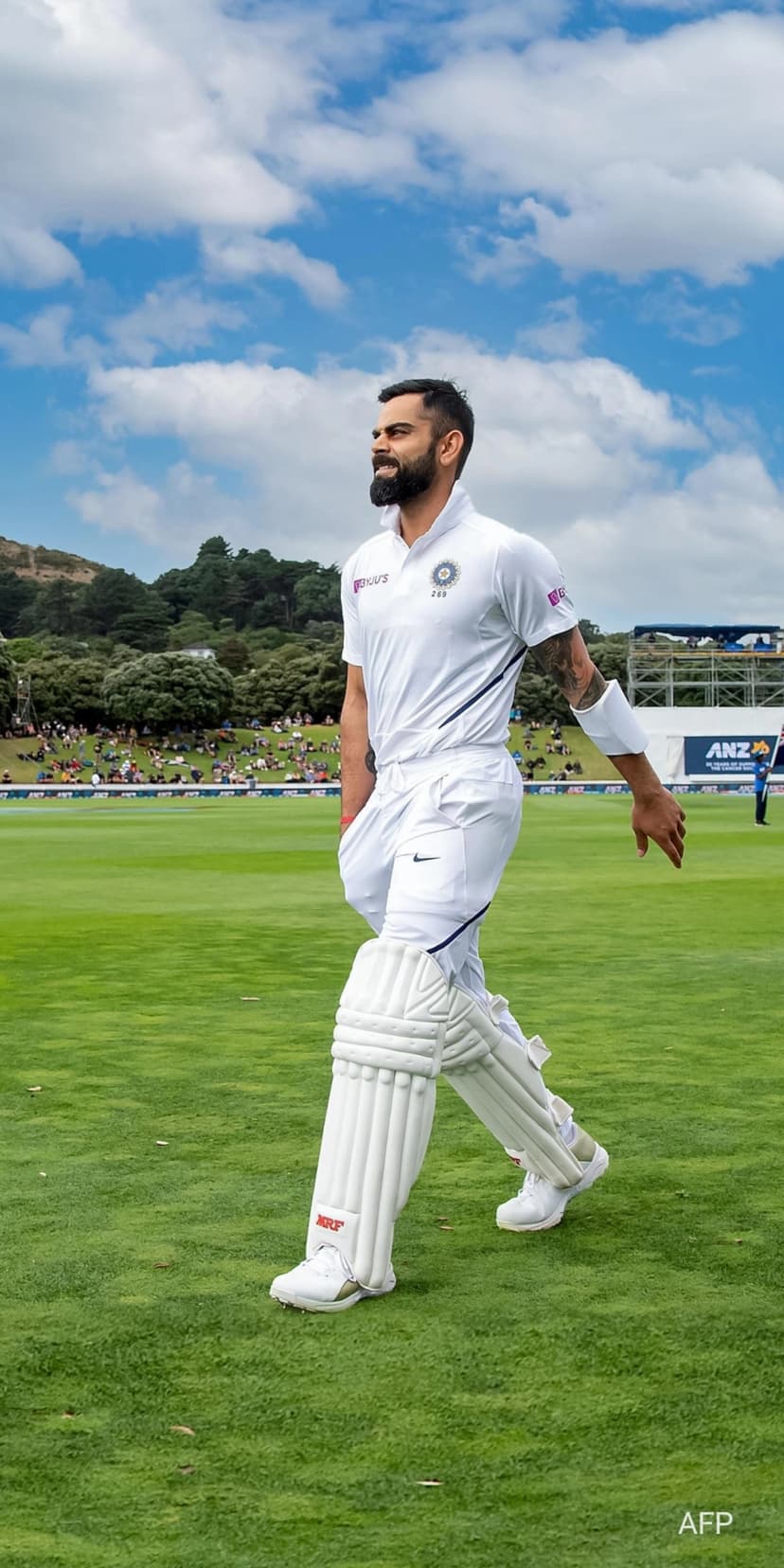 Virat Kohli in white cricket whites and pads walks across a green field under a blue sky with distant hills.