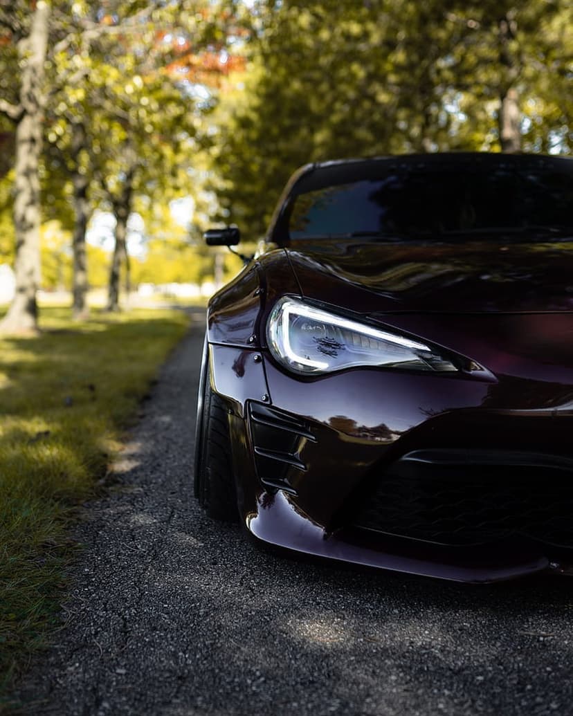 Purple sports car with widebody kit and lit headlights on a gravel road surrounded by blurred forest trees.