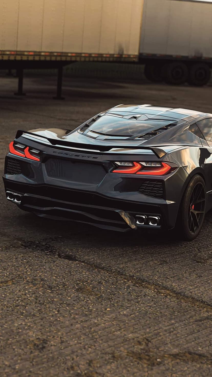 Black Corvette C8 rear view featuring quad exhaust tips and sharp taillights parked on asphalt by a large trailer.