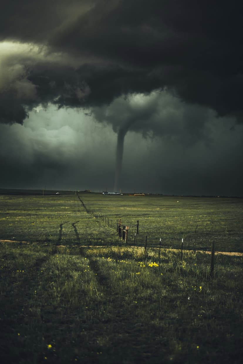 Dark funnel cloud descends onto a grassy field with a wooden fence line under a heavy, swirling storm cell.