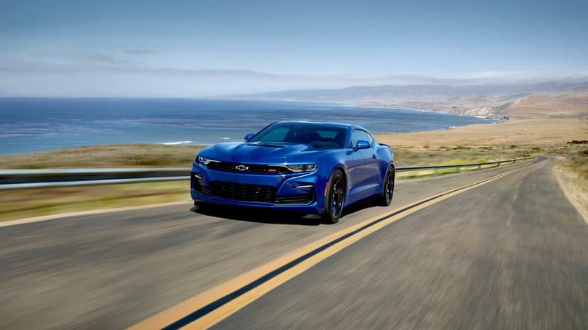 Blue Chevrolet Camaro driving on a coastal highway with ocean views and golden hills under a clear sky.