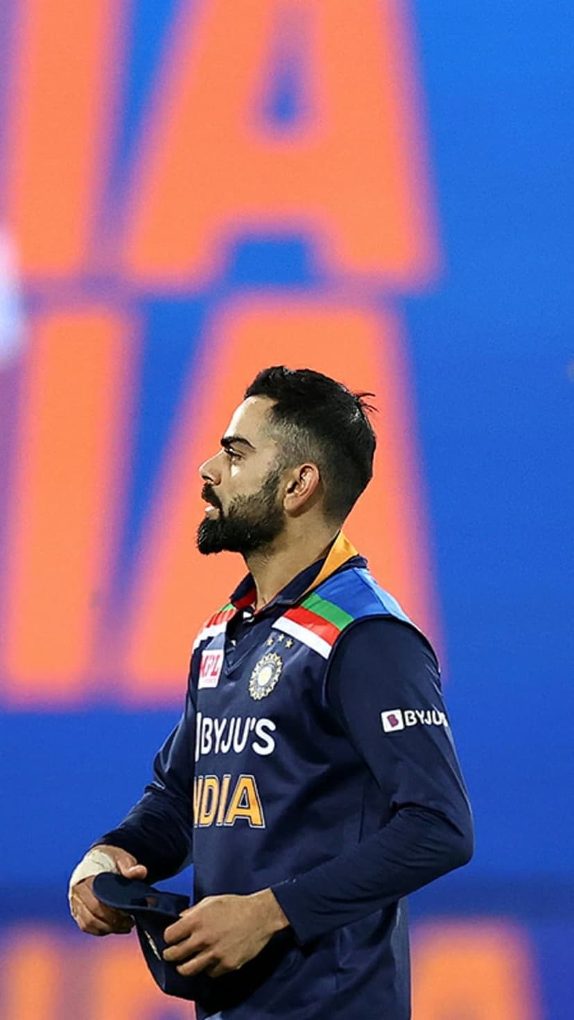 Virat Kohli in a dark blue India jersey stares forward in a profile shot against a blurred stadium background.