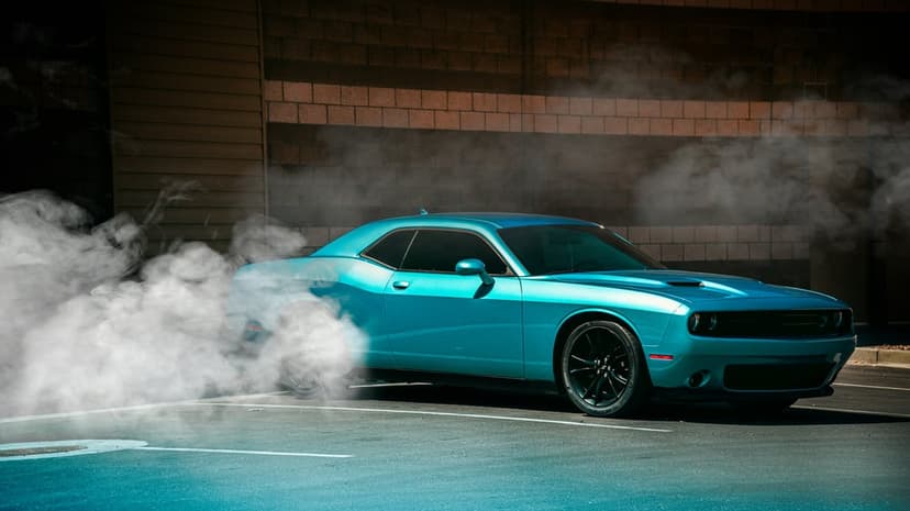 Blue Dodge Challenger performs a burnout with white tire smoke against a dark urban brick wall background.