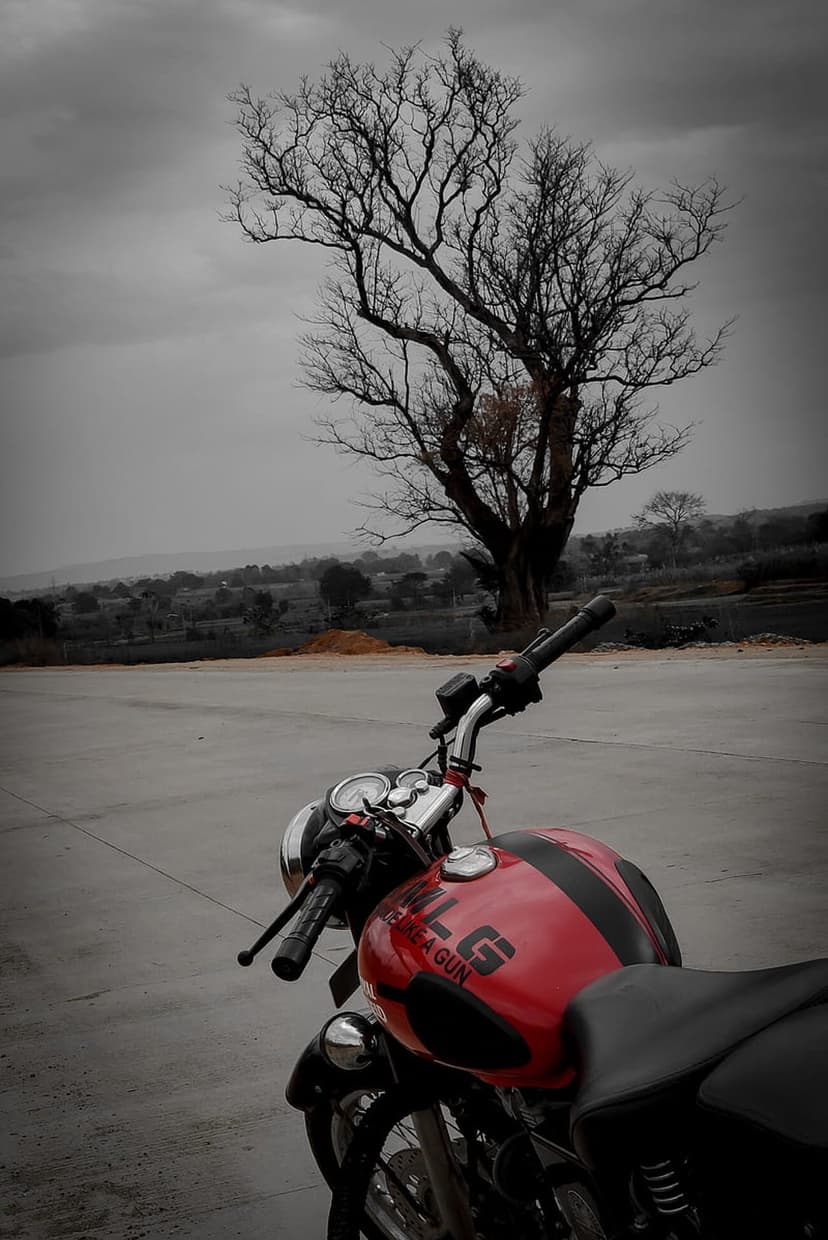 Red motorcycle parked near a bare tree under a cloudy grey sky with rolling hills in the distant background.