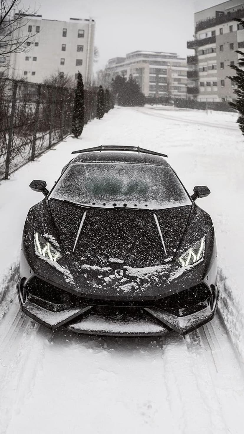 Black Lamborghini supercar with glowing headlights parked on a snowy city street during an active winter snowfall.