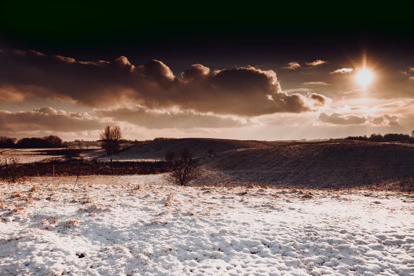 Snow-covered rolling hills under a dark cloudy sky with bright sunlight piercing through the horizon line.