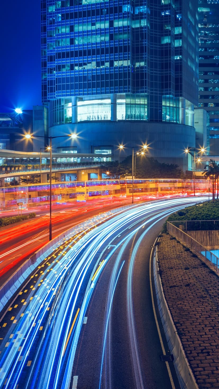 Streaking blue and red light trails flow through a busy city intersection between illuminated skyscrapers at night.