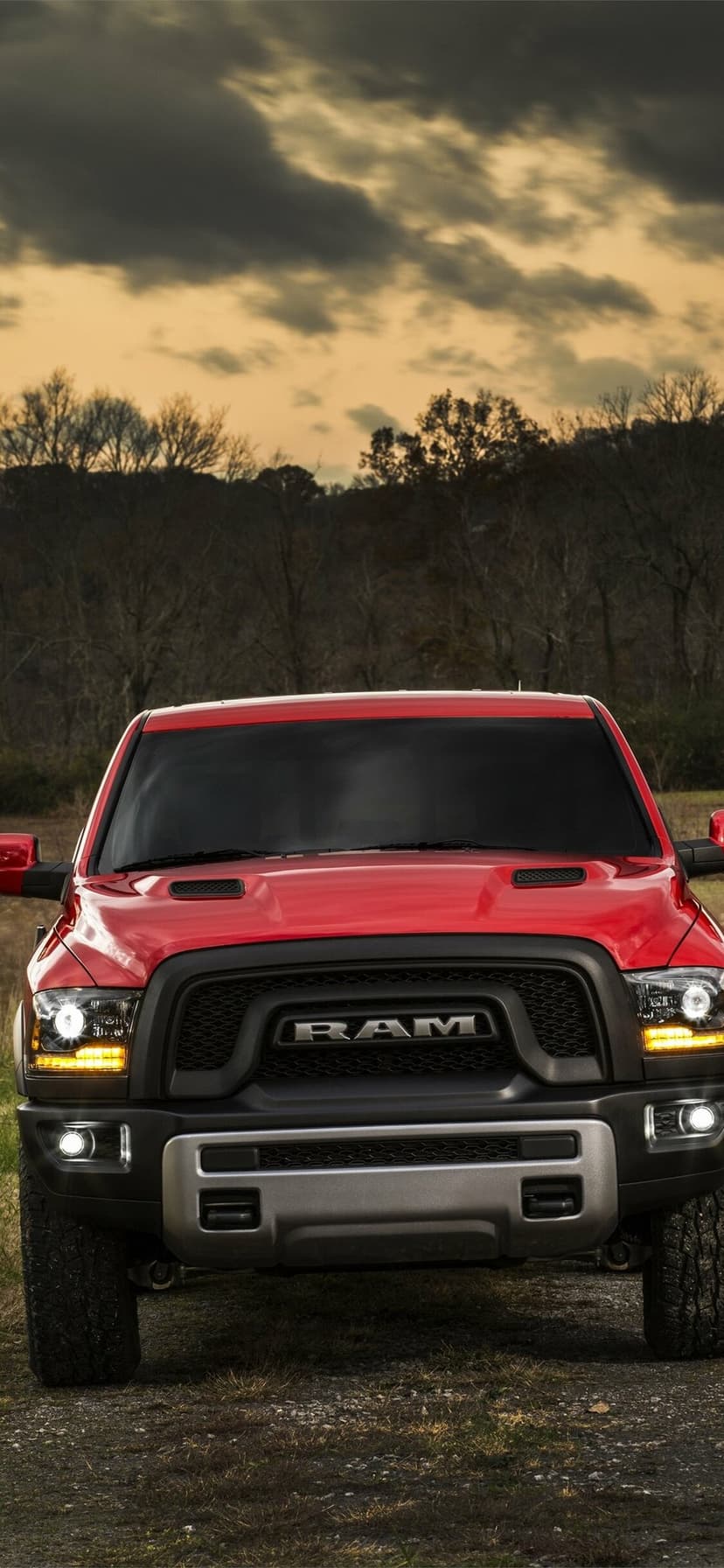Front view of a red RAM truck with illuminated headlights on a dirt path under a cloudy golden sunset sky.