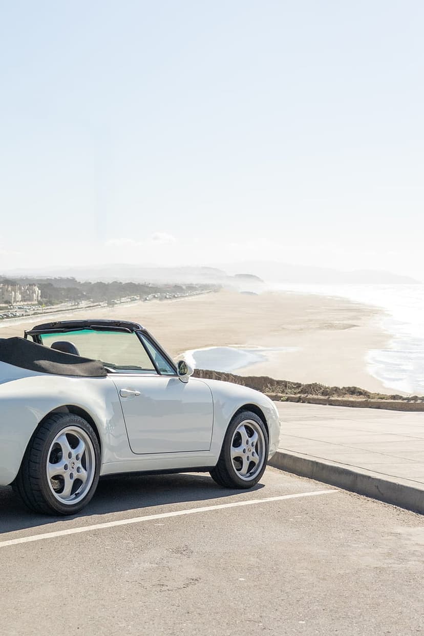 White open-top sports car parked at a cliffside overlook above a sandy bay with turquoise waves and soft skies.