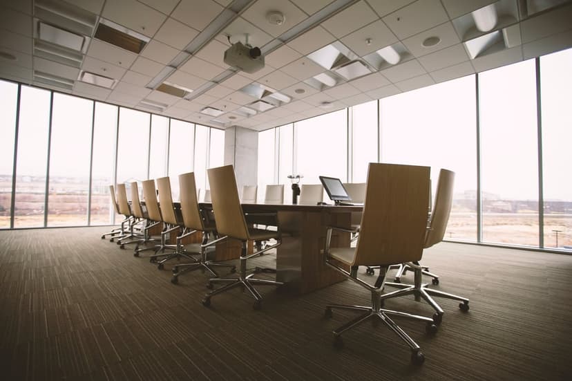 Long wooden conference table and ergonomic chairs in a sunlit office with floor-to-ceiling glass windows.