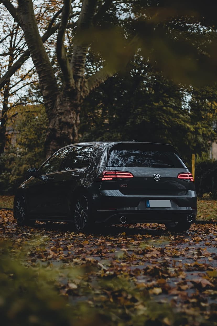 Volkswagen Golf GTI parked on a leaf-strewn forest path with glowing red taillights under a canopy of fall trees.