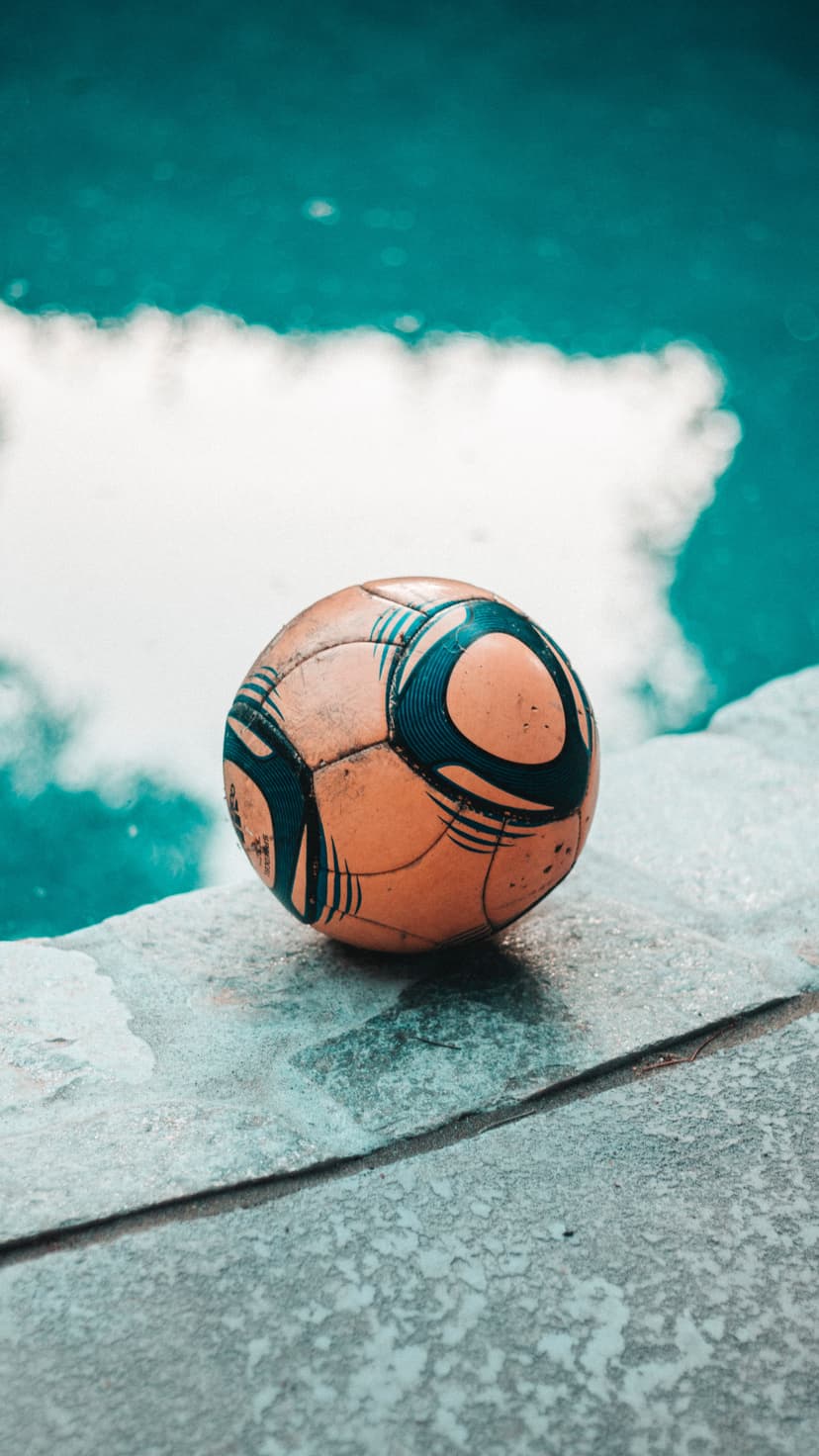 Orange soccer ball with blue panels sits on a concrete pool deck next to bright turquoise water and sky reflections.