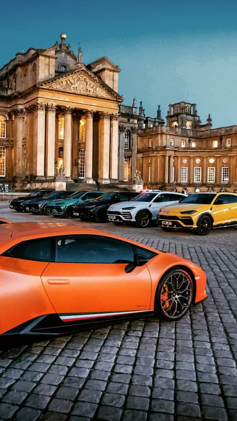 Orange and yellow Lamborghini supercars parked on cobblestone before a lit classical palace at dusk.