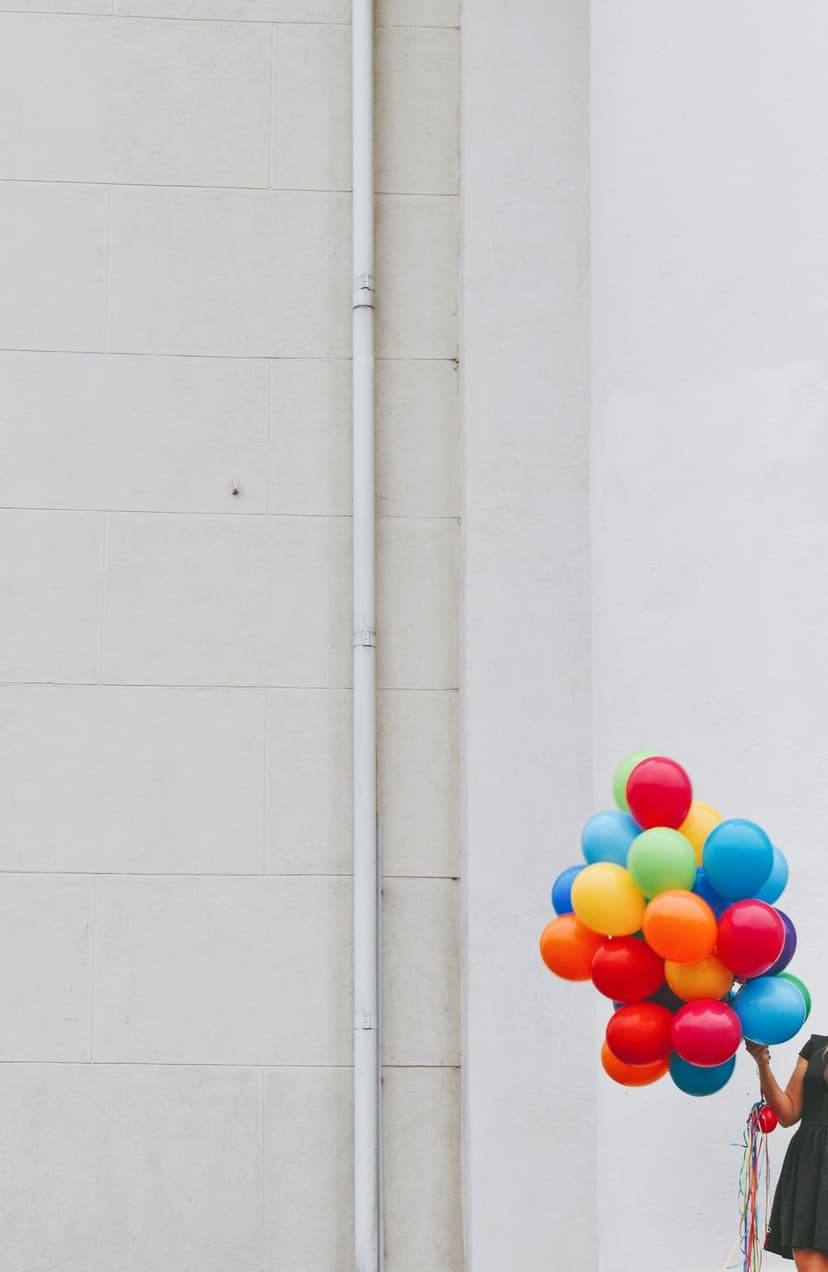 Woman in a black dress holds a bunch of red, yellow, and blue balloons against a white wall with a vertical pipe.