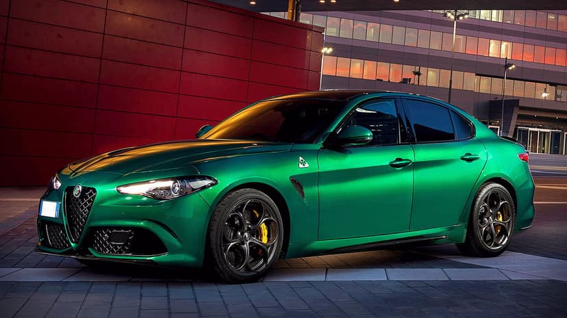 Green Alfa Romeo Giulia Quadrifoglio sports sedan parked beside a red wall and lit office building at dusk.